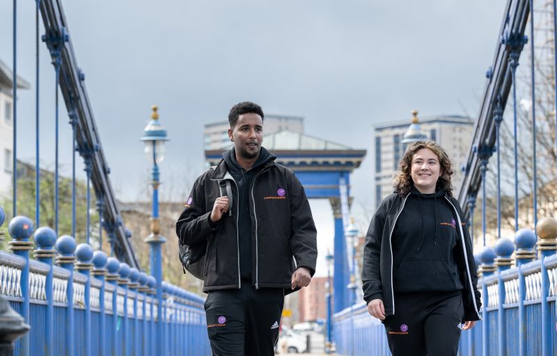 A man and a woman are walking over a blue suspension bridge with tall buildings in the background