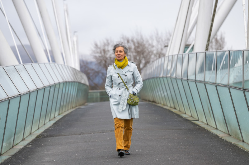 A woman wearing a grey jacket, yellow scarf and brown trousers is walking over a bridge
